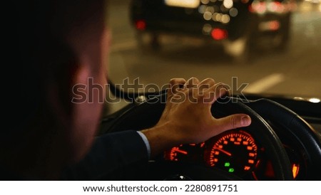 Man driving car fast in night city street with traffic. Road trip, speeding, driver concept. Commuter, male taxi cab driver. Interior back view of male hand on steering wheel in car. Close up Royalty-Free Stock Photo #2280891751