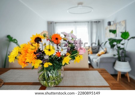 Kitchen counter table with focus on vase with huge multicolor various flower bouquet with blurred background of modern cozy living room with couch and green plants. Open space home interior design.