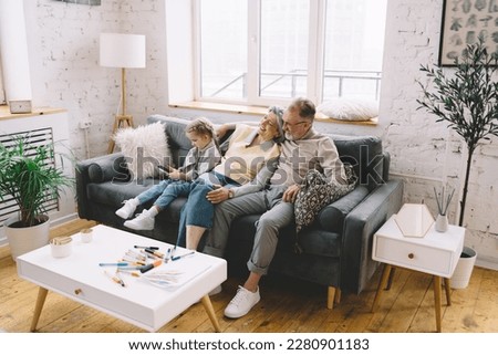 High angle of cheerful senior woman and man resting on cozy couch with cushions in light living room while kid using mobile phone