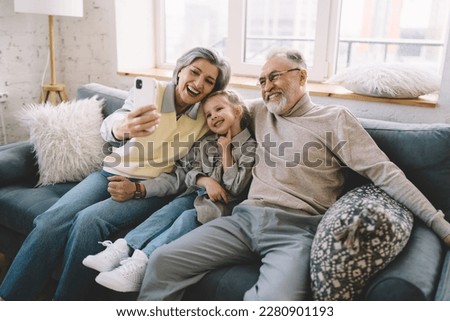 Positive grandparents in domestic clothes sitting together with cute blond girl and taking self portrait while spending time at home