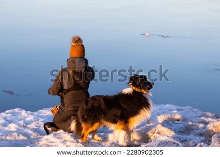 Back view of female photographer kneeling on snow bank waiting for a photo opportunity, with her Bernese Mountain dog on high alert during a winter morning, Quebec City, Quebec, Canada