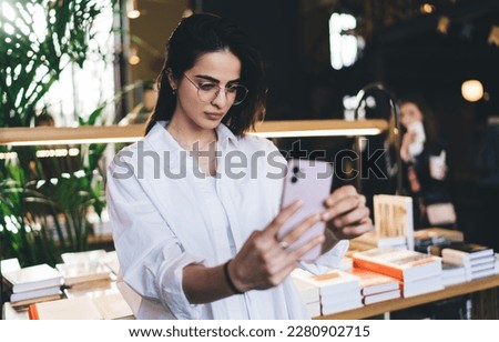 Concentrated female student in eyeglasses looking at screen of mobile phone in hands and taking selfie while standing near neatly arranged publications on table in bookstores for sale