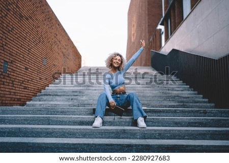 Low angle of happy and cheerful young African American female student with curly hair sitting on steps with hand raised and looking at camera Royalty-Free Stock Photo #2280927683