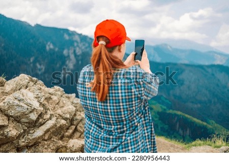 Young woman relaxing on the top mountains, holds a mobile phone in her hands and takes pictures of the landscape. Back view.