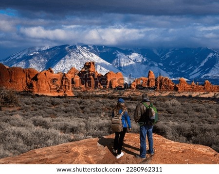 Couple enjoying beautiful mountain view on hiking trip in Utah. The Windows Section of the park, snow covered La Sal Mountains in the background.  Arches National Park ,Moab, Utah, USA. Royalty-Free Stock Photo #2280962311