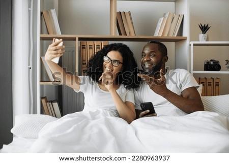 Joyful amorous people getting self-portrait on cell phone while having fun in bedroom at weekend. Smiling multiethnic husband and wife saving precious memories of leisure time together using gadget.