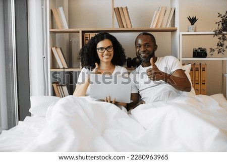 Cheerful multiracial couple holding portable computer and giving thumbs up sign while lying under white blanket at home. Romantic spouses encouraging to living comfortable tech lifestyles.