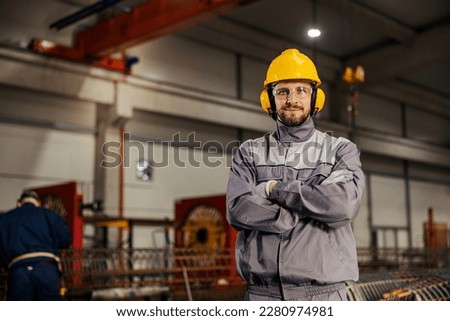 A heavy industry and metallurgy worker in protective uniform is proudly standing in facility and smiling at the camera. Royalty-Free Stock Photo #2280974981
