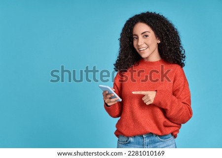 Young happy excited latin woman pointing at mobile phone isolated on blue background. Smiling female model holding cellphone using cell presenting advertising new trendy application concept. Royalty-Free Stock Photo #2281010169