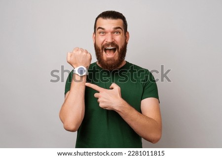 Surprised young bearded man is showing his smart watch and pointing at it over grey background. Royalty-Free Stock Photo #2281011815