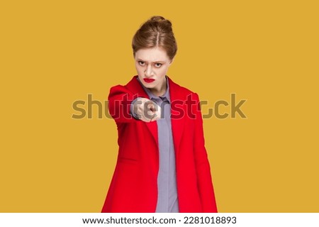 Portrait of strict serious woman with red lips standing indicating index finger to you, looking at camera with bossy expression, wearing red jacket. Indoor studio shot isolated on yellow background. Royalty-Free Stock Photo #2281018893