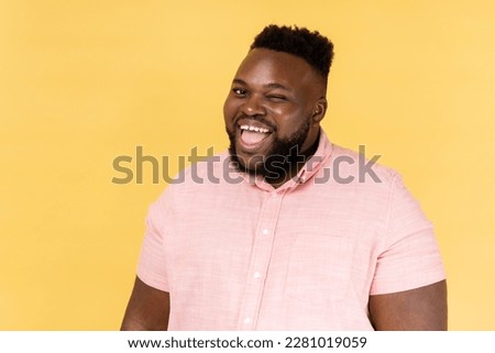 Portrait of childish happy young adult man wearing pink shirt winking and optimistically smiling, good mood, cunning bearded guy flirting. Indoor studio shot isolated on yellow background. Royalty-Free Stock Photo #2281019059