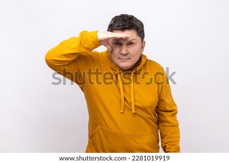 Thoughtful dark haired man looking far away at distance with hand over head, attentively searching for bright future, wearing urban style hoodie. Indoor studio shot isolated on white background. Royalty-Free Stock Photo #2281019919