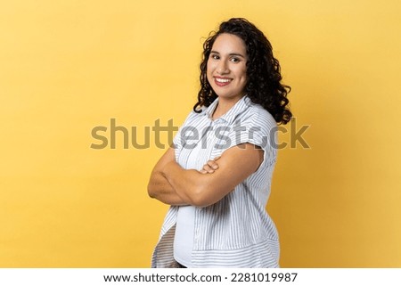 Portrait of satisfied delighted happy woman with dark wavy hair standing with folded hands and looking at camera with toothy smile. Indoor studio shot isolated on yellow background. Royalty-Free Stock Photo #2281019987