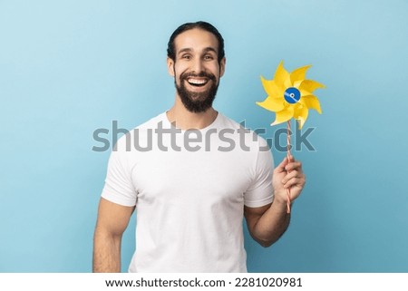 Portrait of cheerful childish man with beard wearing white T-shirt holding paper windmill, pinwheel toy on stick, playing with paper toy. Indoor studio shot isolated on blue background. Royalty-Free Stock Photo #2281020981
