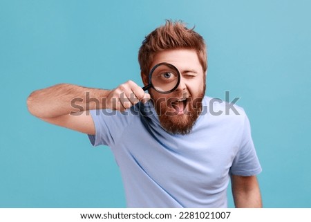 Portrait of funny positive bearded man holding magnifying glass and looking at camera with big zoom eye, curious face, keeps mouth open. Indoor studio shot isolated on blue background.