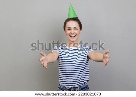 Come into my arms. Portrait of pleased smiling woman wearing striped T-shirt and party cone, inviting her friends to birthday party. Indoor studio shot isolated on gray background.