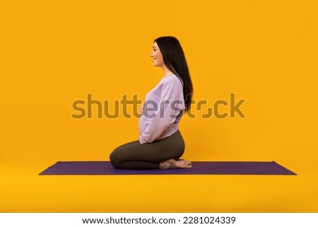 Prenatal yoga. Pregnant lady sitting on fitness mat and touching belly, enjoying pregnancy time, ready for training on yellow studio background, side view