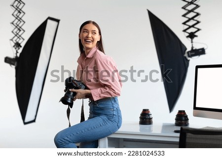 Excited female photographer sitting on edge of table, holding her DSLR camera, looking aside and smiling, working with computer in modern photostudio interior
