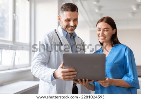 Happy male doctor and female nurse talking, working together, using laptop, discussing patient medical checkup results or diagnosis, wearing uniform holding electronic device
