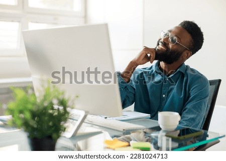 Optimistic dream. Happy black male employee sitting at workplace in front of computer and smiling, visualizing future career growth. Male enterpreneur thinking on good business decision Royalty-Free Stock Photo #2281024713