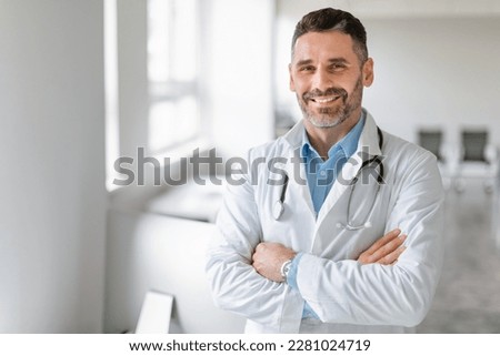 Portrait of friendly middle aged european male doctor in workwear with stethoscope on neck posing with folded arms in clinic interior, looking and smiling at camera, free space Royalty-Free Stock Photo #2281024719