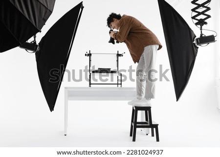 Professional male photographer standing on ladder and taking high angle content photos for modern shop, working at the professional photostudio, side view