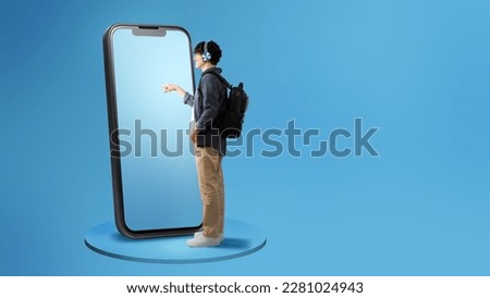 Side View Of Guy Using Large Cellphone Touching Screen Wearing Headphones Standing With Backpack Over Blue Studio Background. Panorama With Free Space For Text, Full Length