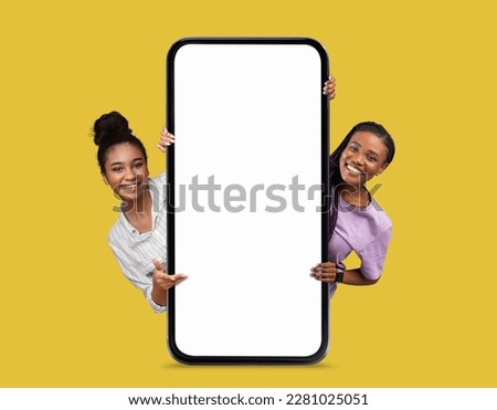 Online Offer. Two Beautiful Smiling Black Women Peeking Out Behind Big Blank Smartphone With White Screen Isolated Over Yellow Background, African American Females Showing Copy Space, Collage, Mockup Royalty-Free Stock Photo #2281025051