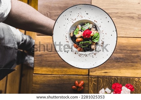 Unrecognisable waiter serving black tagliolini pasta dish with shrimp and tomatoes on white minimalist plate. Italian cuisine. Restaurant food. Horizontal indoor top-down shot. High quality photo