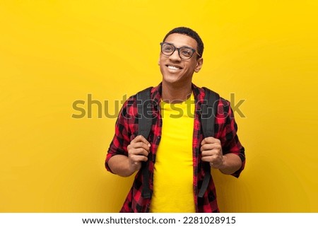 young guy african american student in glasses with backpack looks at copy space on yellow isolated background, student latino in shirt, concept of learning and education