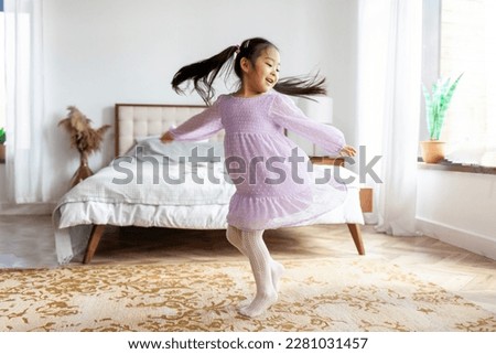 little Asian girl in dress dances and spins at home, Korean child in festive outfit moves and rejoices in the room near the bed