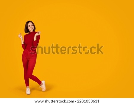Excited curious smiling beautiful brunette young woman wearing red casual outfit looking at copy space for advertisement and snap fingers, orange studio background, exciting offer, full length