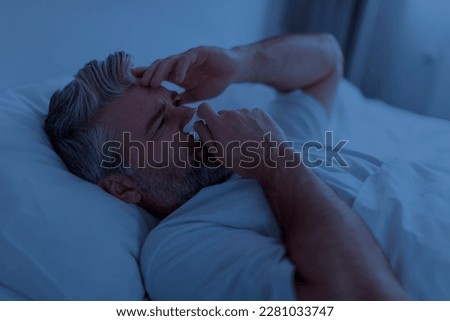 Closeup shot of sick middle aged grey-haired man wearing pajamas touching runny nose with napkin, sneezing in bed at night, touching his head, suffering from cold, flu, coronavirus Royalty-Free Stock Photo #2281033747