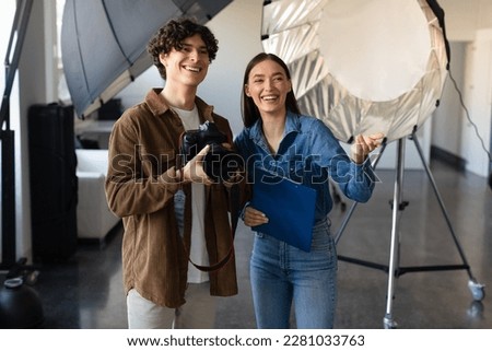 Young male photographer talking with female art director, talking and preparing for photo shooting, woman pointing forwards and smiling, standing near modern lighting equipment Royalty-Free Stock Photo #2281033763