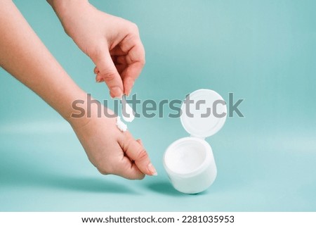 Face cream in a white jar on a blue background. Young woman holds cream in her hands. Closeup Skincare Product for facial. Woman applying cream on her hand