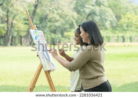 Asian mother and cute African daughter smiling holding same paintbrush and painting coloring watercolor on canvas together in green park on summer. Mom support little artist girl in outdoor activity