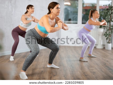 Concentrated young girl doing yoga with group of women in modern studio, performing horse yoga pose to strengthen muscles of legs and buttocks. Bodyweight training concept.. Royalty-Free Stock Photo #2281039805