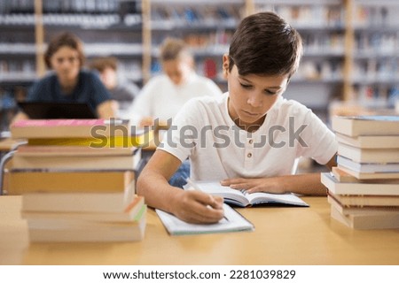 Young boy sitting at table in reading room in library and doing homework.