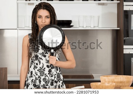 Young woman poses as a warrior armed with a frying pan in the kitchen. Ironic image inspired by past advertisements. She has box braids, the kitchen is modern and bright, and she is beautiful