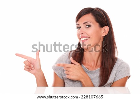 Portrait of attractive brunette on grey t-shirt pointing to her right while smiling at you on isolated white background - copyspace