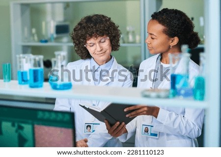 Young brunette female researcher in lab coat and eyeglasses looking at notes in notebook held by African American colleague Royalty-Free Stock Photo #2281110123