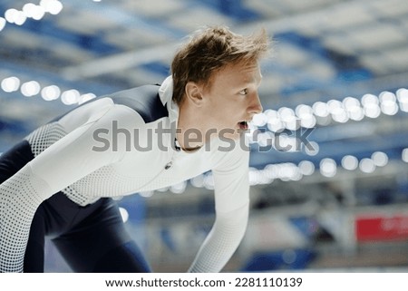 Youthful blond male athlete in sports uniform waiting for start signal while standing on skating rink and looking aside during competition