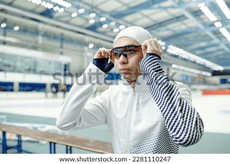 Youthful guy in sports uniform sitting in front of camera at break and putting on eyeglasses before moving to ice skating arena