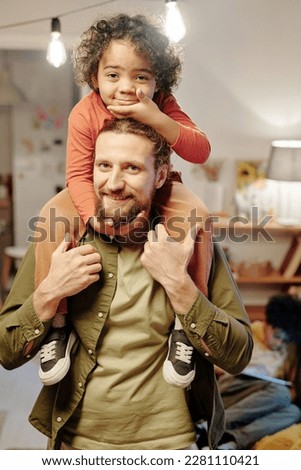 Happy young man looking at camera while standing in living room and holding his adorable little son on shoulders while having fun