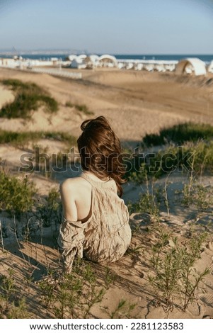 red-haired woman posing sitting with her back to the camera in a beige dress on the sea coast during sunset