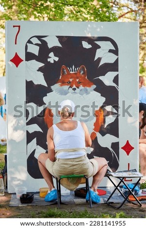 Girl artist draws with paint brush surreal animal playing card on white canvas at outdoor art painting festival, paintings art picture process, rear view. Woman artist paints atmospheric picture