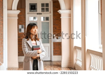 female Asian research student wearing stylish grey overcoat holding a book in university campus in Hong Kong Royalty-Free Stock Photo #2281150223