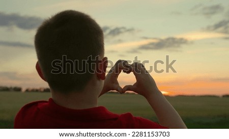 Child boy plays sun in park, looks through symbol of heart at sky. Family love. Child makes heart shape with his fingers against sunset. Childrens hands form heart symbol. Love, Health. Kid dream, Sky
