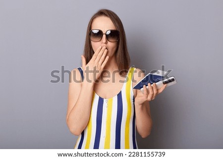 Photo of shocked astonished woman tourist holding cellphone and passport covering mouth with palm booking wrong day for departure wearing striped swimming suit isolated over gray background